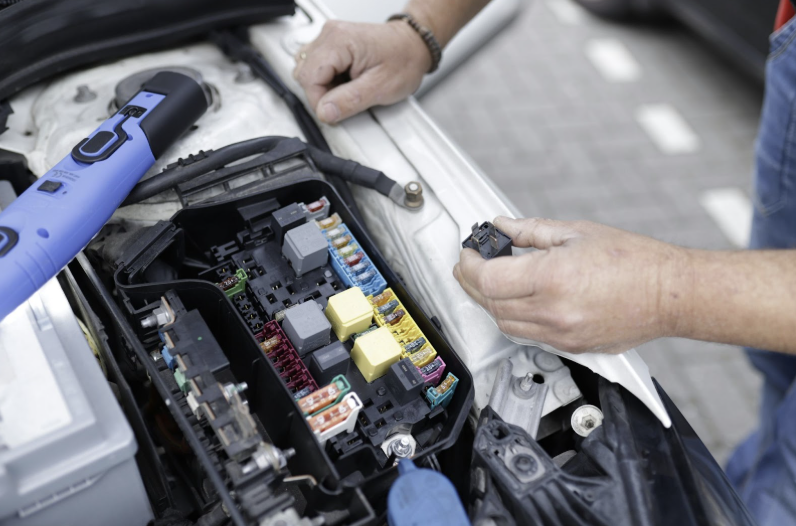 Mechanic holding a relay while inspecting the car’s open fuse box in the engine bay, with a diagnostic tool nearby.