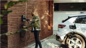 A man plugs a cable into a wall-mounted EV charger outside a modern wood-paneled home.